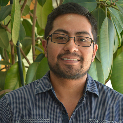 Omar Gomez is wearing a checkered shirt and standing front of a plant.