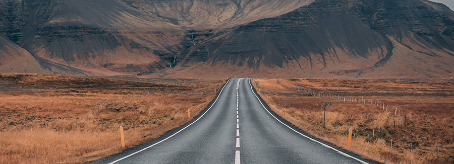 Image facing a long highway road surrounding by rust-colored fields leading to a mountain.