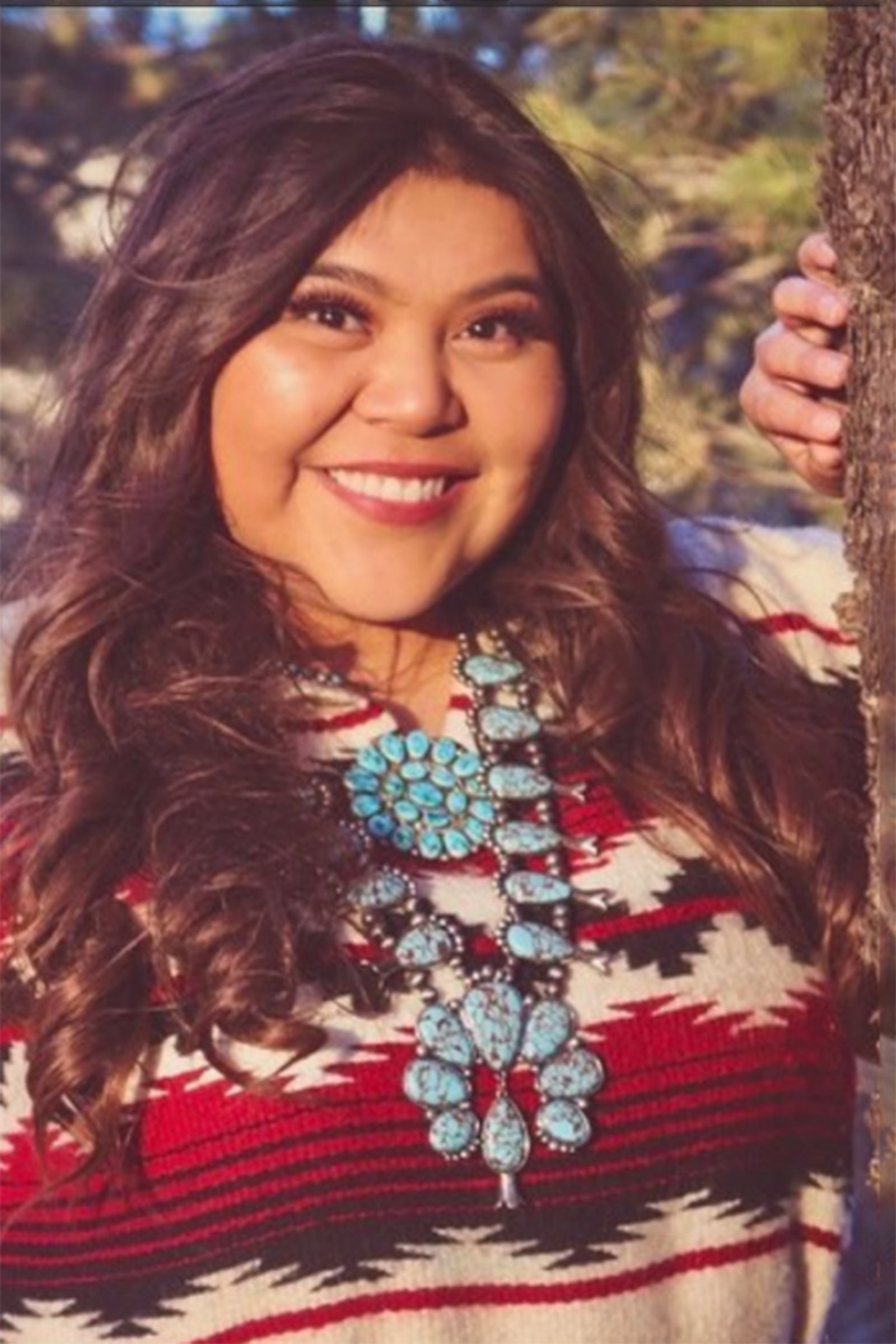 Stacey Litson standing by a tree wearing traditional clothing and wearing Native American jewelry.