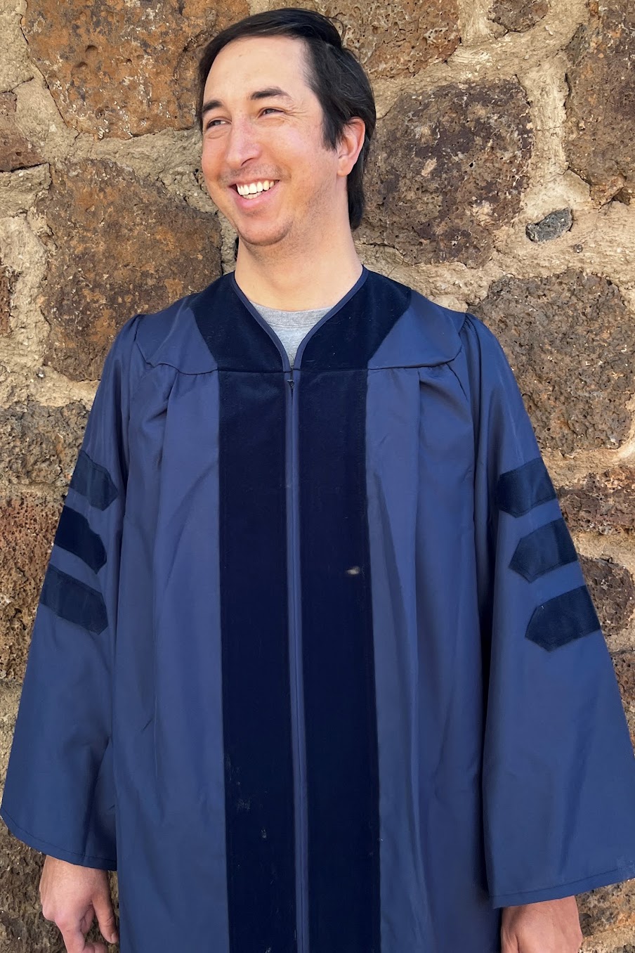  Travis Pinn wearing graduate attire standing in front of outdoor brick wall.