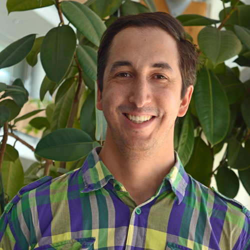 Travis is wearing a multiple colored shirt and standing in front of a large plant.