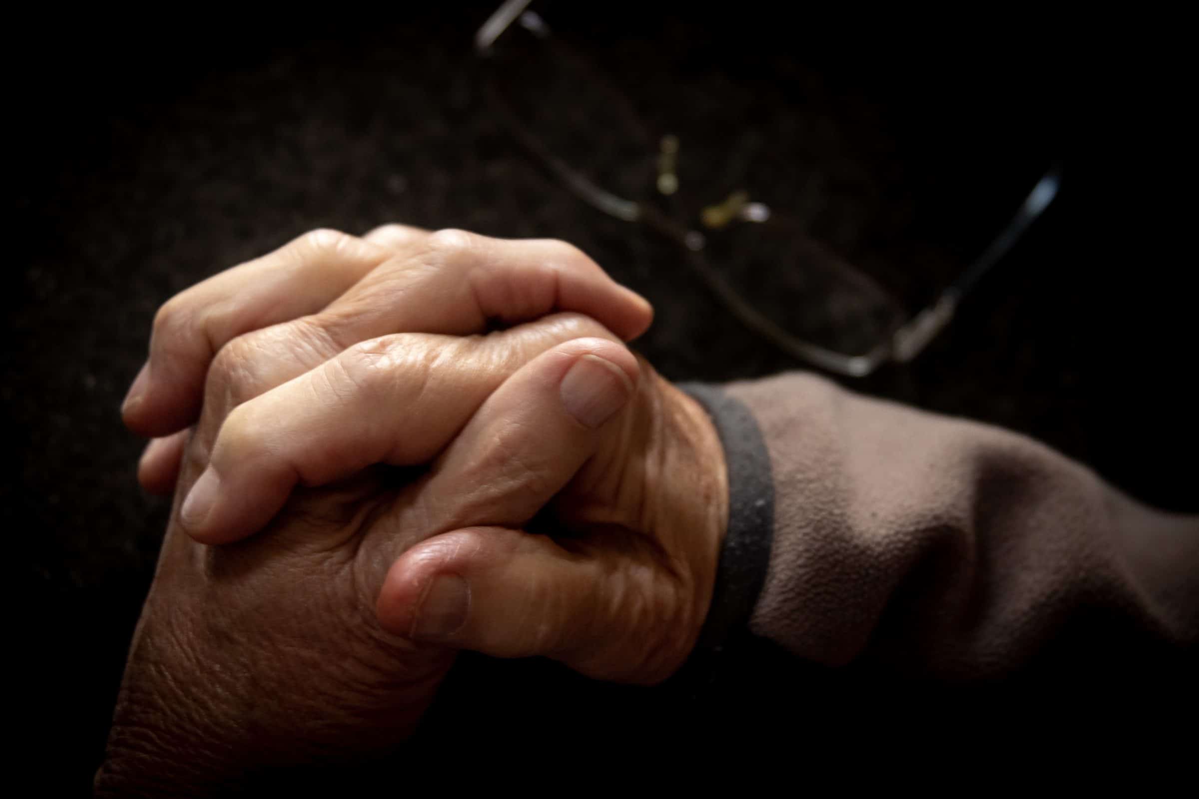 Wrinkled and aged hands clasped together against a dark background.