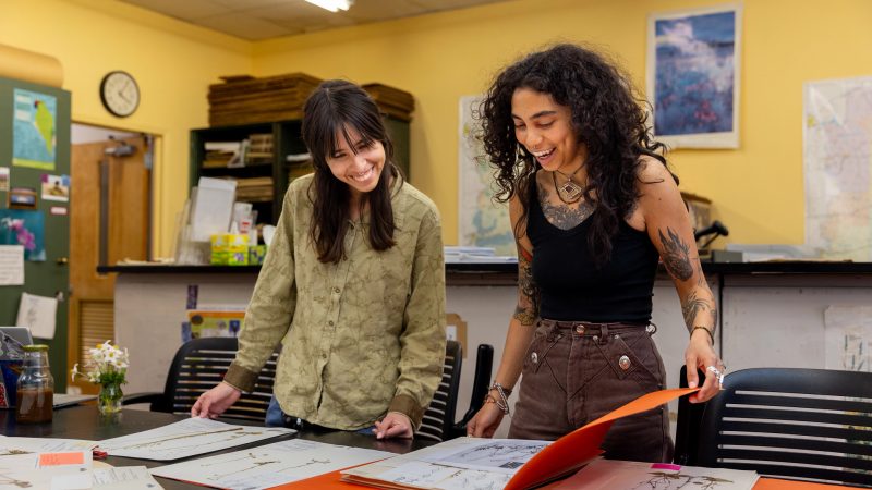 Two students laughing and talking about a book with environmental images inside.