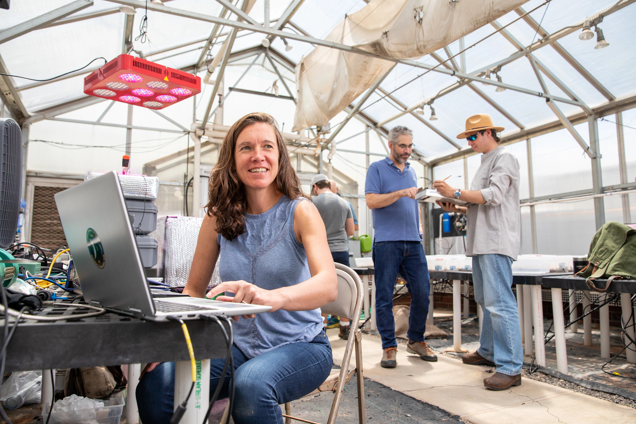 A women working on her computer and sitting at a desk in a greenhouse.