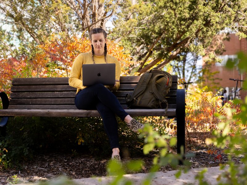 A women sitting on a bench outdoors while working on her computer.