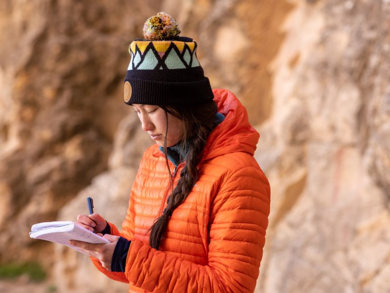 A student wearing an orange jacket and beanie while writing on a note pad at the Grand Canyon.