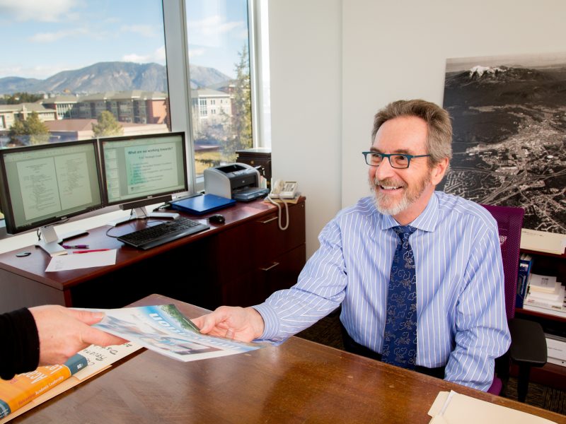 Dean Jagodzinski sitting at his desk in an office, handing a pamphlet to a visitor.