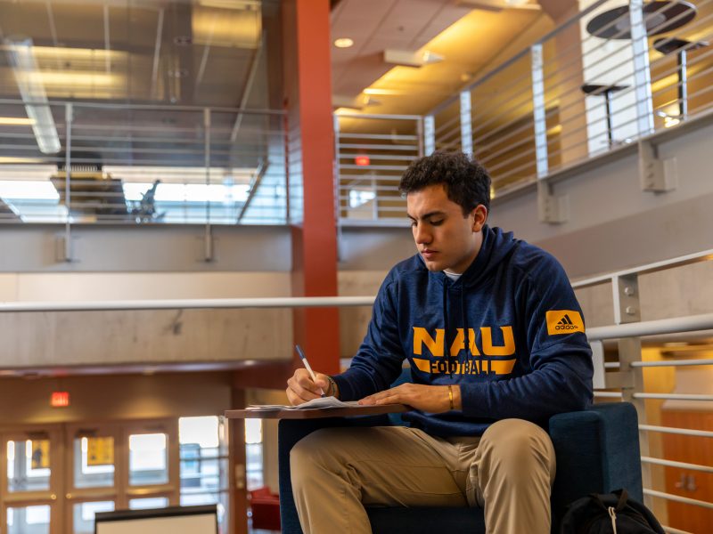 A student wearing a N A U hoodie, sitting down writing something down.