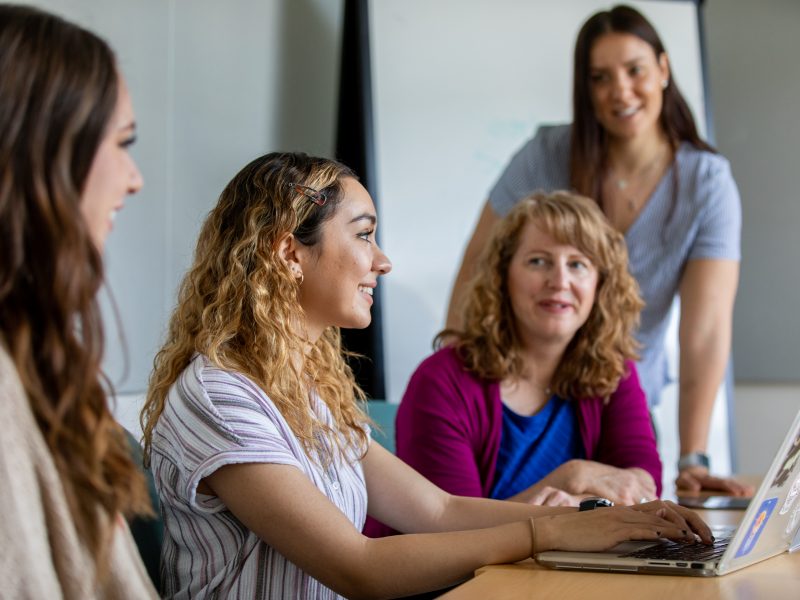 Four women sitting at a table, with one using a laptop.