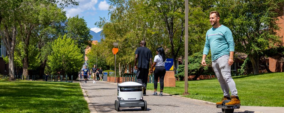 Campus pedway in the summer with a starship robot, a student on a one wheel and a couple walking in the distance. 