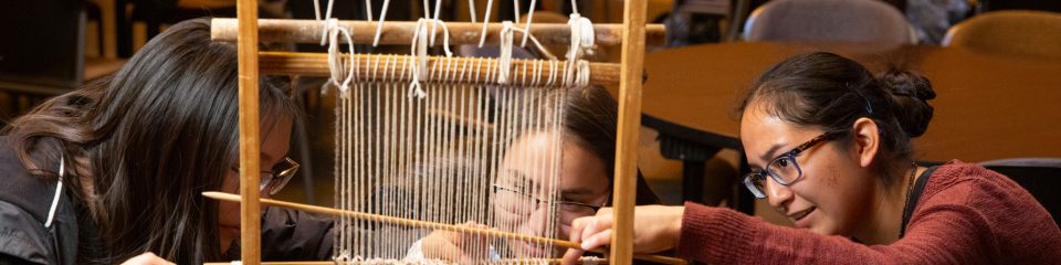 Three indigenous females learning to use a traditional loom