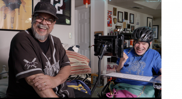 a smiling man sitting next to another smiling man who is wearing a head pointing device