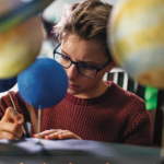 a student sits at a desk surrounded by model planets and looking concentrated