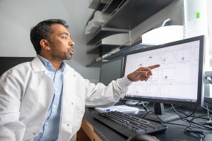 Narendiran Rajasekaran points at his computer screen. He is wearing a white lab coat and has short dark and gray hair.
