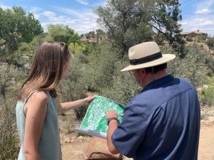 Catrin Edgeley holds a map with an unidentified man who is pointing to a place on the map. They are standing in front of a group of trees in the Southwest with sand around..