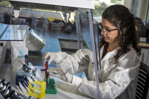 Emily Cope, PhD, in her lab looking down at test tubes she is holding. She is wearing a white lab coat and has brown, curly hair pulled back in a clip and she is wearing glasses