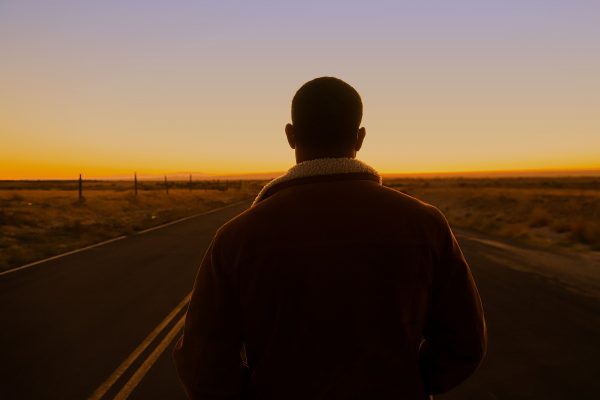 Man in jacket with fur collar stands on a road looking into sunset over mountains in the distance