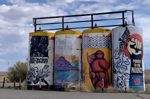 Barrels on the Navajo Nation that are painted with graffiti art, which includes birds and an arm pointed vertically with a hand in a fist that says "Power to the Patients."