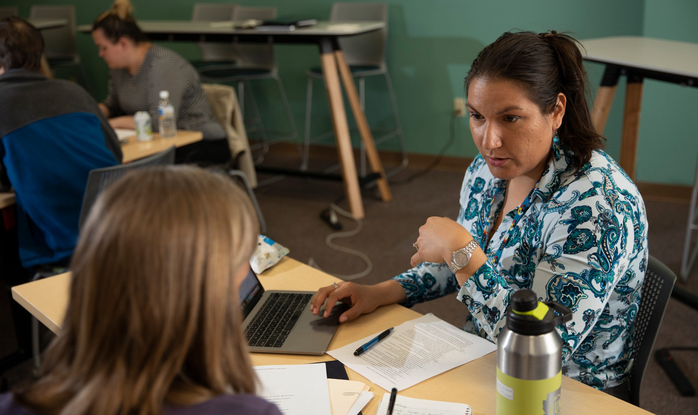 An NAU professor teaches a student manuscript writing. They are both sitting at a table, and the professor is in front of a laptop. She has dark hair pulled into a ponytail and is wearing turquoise earrings and a white button-down shirt with turquoise paisley print. The student has their back to the camera, but has straight, light brown hair.