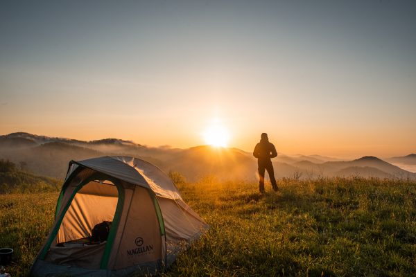 Man in silhouette looks at the sunset over the mountains with tent in foreground