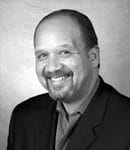A black and white headshot of Rick Rodriguez, a man with a beard wearing a dark blazer and lighter shirt