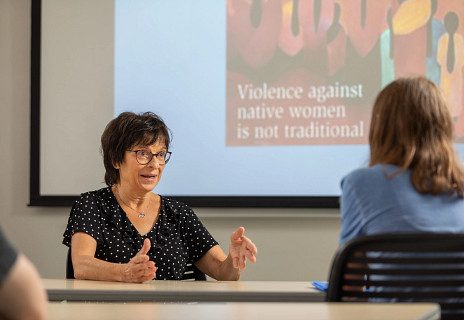 Faculty member and student interacting in the classroom in front of projector screen that reads, "Violence against native women is not traditional"